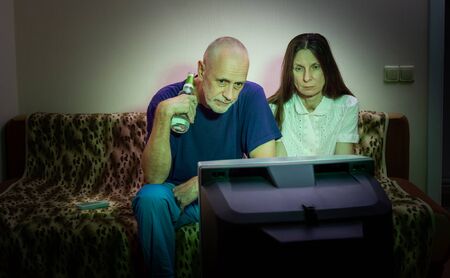 Middle-aged man and woman, watch television, looking sad, sitting on a couch. They watch a sad movie or listen to bad news that makes them a little unhappy and depressedの写真素材