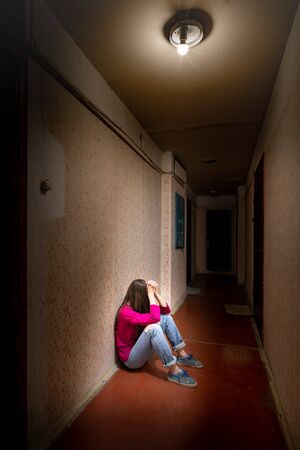 A sad and desperate woman sits in a dark corridor illuminated by a gloomy light. Her pain and her many problems pushed her into complete isolation. His sadness is only equalled by his loneliness.の写真素材