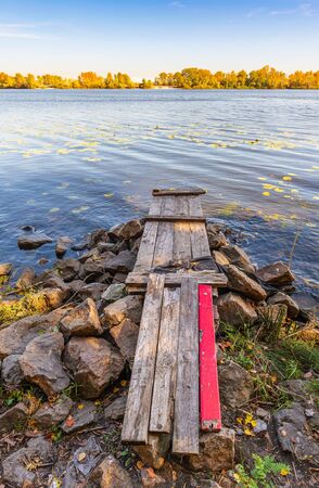 A wooden pontoon with a red paint plank  over big stones close to the blue Dnieper river is waiting for the fisherman.の写真素材