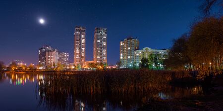 Night view of modern buildings in the obolon district of Kiev, Ukraine, viewed from the southern part of Natalka Park. The moon shines in the starry blue sky. The lights reflect on Dnieper river.の写真素材