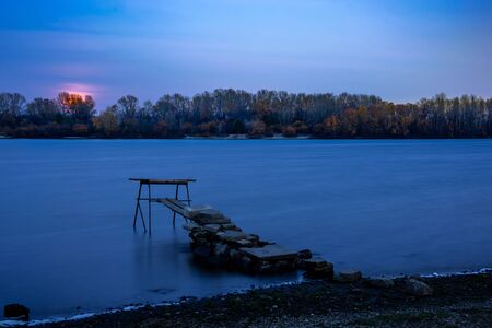 Moon through the autumn forest close to Dnieper river in Kiev, Ukraine. Soft clouds in the dark sky cover partially our natural satellite. The water looks like a glossy blue mirror.の写真素材