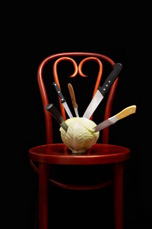 Symbolic conceptual image of a white cabbage on a wooden Thonet chair. Knives are stuck into the vegetable and symbolize cooking, violence, crime, suffering.  On black background.の写真素材
