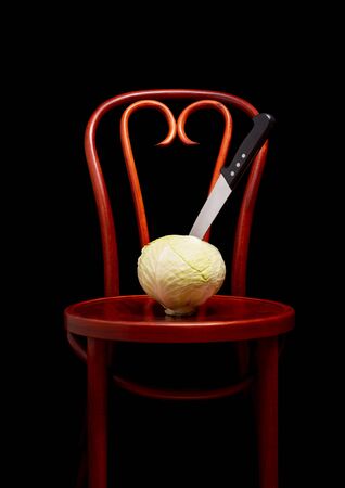 Symbolic conceptual image of a white cabbage on a wooden Thonet chair. A knife is stuck into the vegetable and symbolize cooking, violence, crime, suffering.  On black background.の写真素材