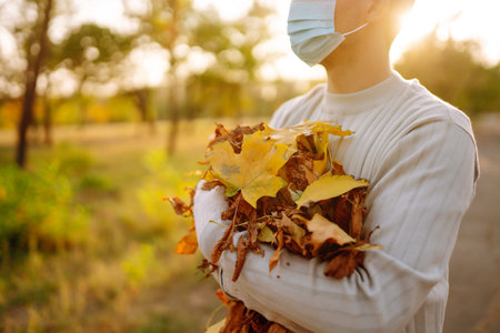 Man in protective medical mask cleans autumn leaves in the park. Man in gloves collects.の写真素材