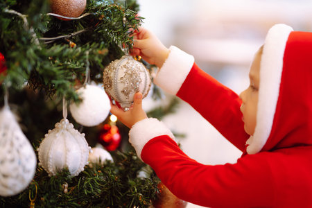 Little child hanging decorative toy ball on Christmas tree branch. Cute child in santa costume decorating.の写真素材