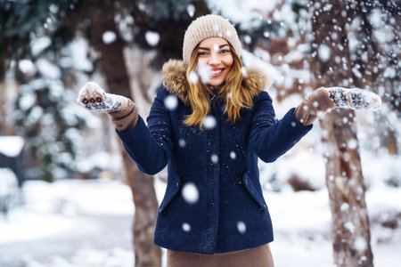 Beautiful girl playing with snow in winter forest. Smiling girl in a blue jacket and knitted hat.の写真素材
