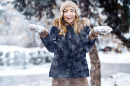 Beautiful girl playing with snow in winter forest. Smiling girl in a blue jacket and knitted hat.の写真素材