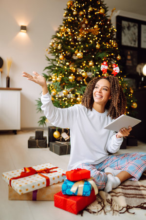 A young woman on the background of a Christmas tree with gifts, with a tablet has a video call or video chat.の写真素材