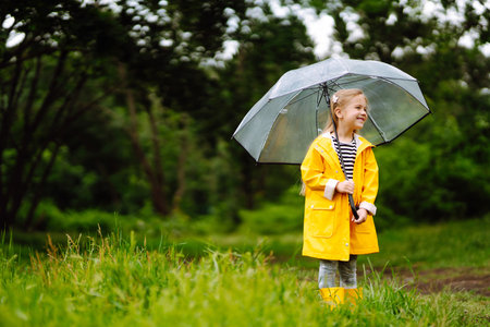 Little girl with transparent umbrella playing in the rain on sunny. Beautiful girl in a yellow cloak and rubber boots.の写真素材