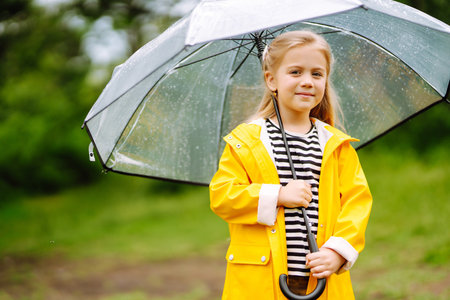 Little girl with transparent umbrella playing in the rain on sunny. Beautiful girl in a yellow cloak and rubber boots.の写真素材