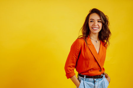 Portrait of young girl posing on yellow background. Pretty woman wears a carrot blouse and jeans.の写真素材