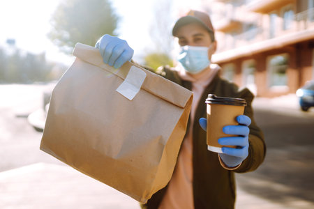 Courier in protective mask and medical gloves delivers takeaway food and coffee. Covid-19. Stay at...