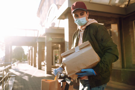 Courier in medical mask and gloves delivers on a scooter a bag of food in quarantine city....