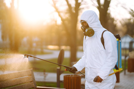 Man in protective suit and mask sprays disinfector onto the railing in the public place. Covid-19.の写真素材