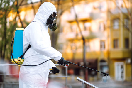 Man in protective suit and mask sprays disinfector onto the railing in the public place. Covid-19.の写真素材