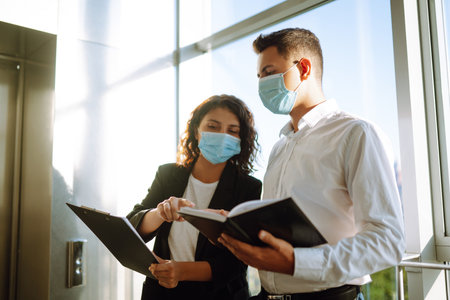 Two coworkers in protective face masks discussing project. Work in office during quarantine. covidの写真素材