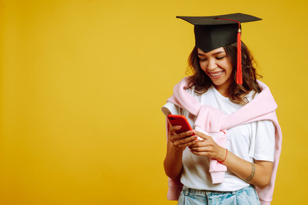Graduate woman in a graduation hat take selfie on a yellow background. African American woman with phone.の写真素材