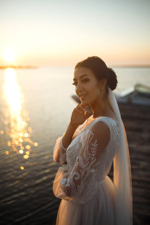 Beautiful bride in an elegant white dress on a bridge against the background of the sea and in the rays.の写真素材