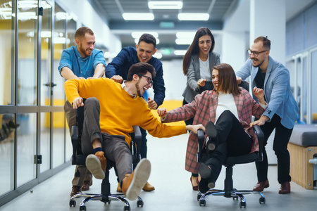 Young cheerful business people racing on office chairs and laughing. Funny activity