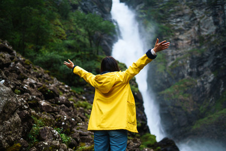 Girl tourist in yellow jacket enjoys freedom against backdrop of waterfall. Traveling, lifestyle.