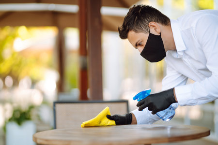 Young Waiter in protective face mask and gloves sanitizing. cleaning the table with disinfectant