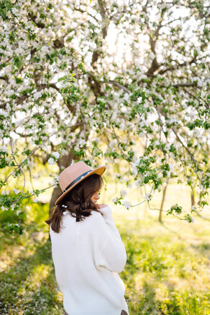 Portrait of Woman in hat posing near flowering tree. Smiling young woman enjoying the smell of flowers.の写真素材
