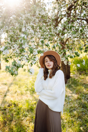 Portrait of Woman in hat posing near flowering tree. Smiling young woman enjoying the smell of...