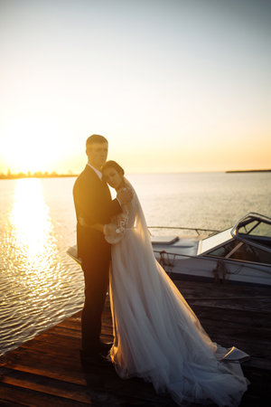 Beautiful bride and stylish groom together on the bridge against the background of the boat at sunset.の写真素材