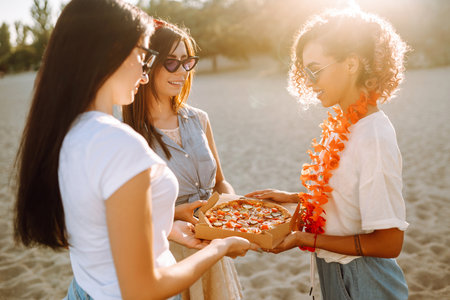Three young woman spending time together at the beach, having a picnic, eating pizza.の写真素材