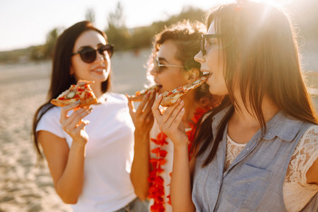 Three young woman spending time together at the beach, having a picnic, eating pizza.の写真素材