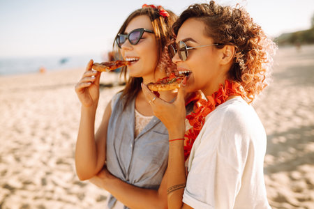 Two young women resting together on the beach and eating pizza. fast food concept. beach holidays.の写真素材