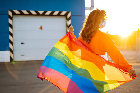 African American lesbian woman with medical face mask posing with lgbt pride flag. Curly woman during quarantine of coronavirus pandemic.の写真素材