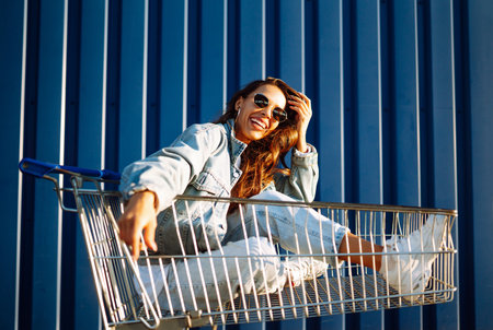 Young woman with phone sitting shopping cart having fun near color wall. Shopaholic, customer.の写真素材