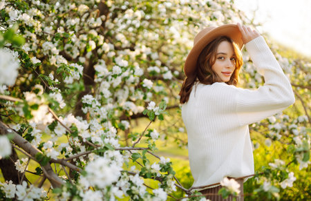Portrait of Woman in hat posing near flowering tree. Smiling young woman enjoying the smell of flowers.の写真素材