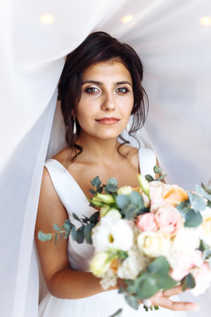 Young bride in white dress with a bouquet of flowers posing under curtain. Morning of the bride.の写真素材