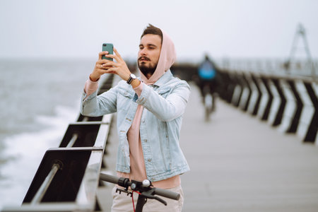 Young man with phone riding electric scooter on a pier near sea. Ecological transportation concept.の写真素材