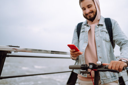 Young man with phone riding electric scooter on a pier near sea. Ecological transportation concept.の写真素材