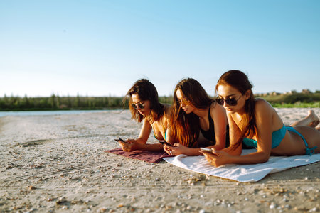 Three beautiful girls using mobile smart phone at the beach. Girls sunbathing on the beach.の写真素材