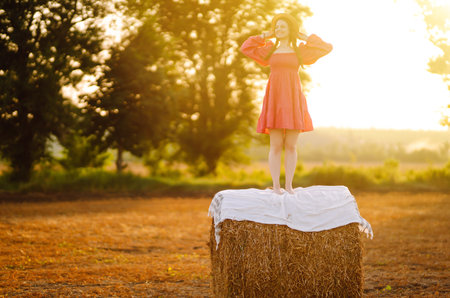 Beautiful curly woman in hat and clothes posing near hay bales in the countryside at sunset.