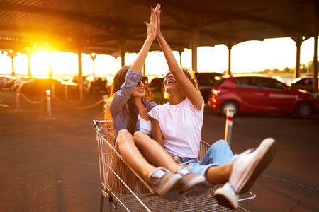 selfie time! Two beautiful girls having fun on shopping trolleys. Multiethnic young people racing on shopping cart.の写真素材