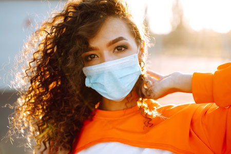 Young woman with medical face mask at sunset. Curly woman posing during quarantine of coronavirus pandemic.の写真素材