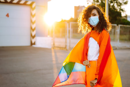 African American lesbian woman with medical face mask posing with lgbt pride flag. Curly woman during quarantine of coronavirus pandemic.の写真素材