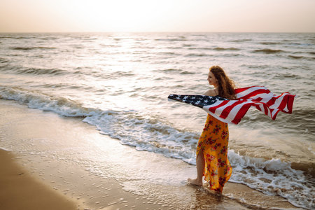 Young woman with american flag on the beach at sunset. 4th of July. independence day.の写真素材