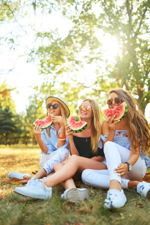 Three nice girls having fun eating watermelon In the park. Excellent sunny weather. Beautiful figuresの写真素材