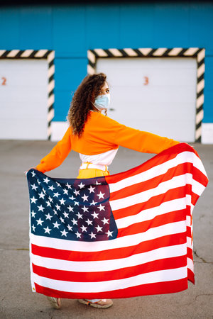 African American woman with medical face mask posing with american flag.The concept of preventing the spread of the epidemic. Covid-2019.の写真素材