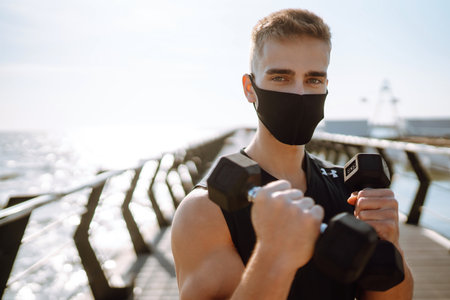 Handsome muscular man in protective mask, sports outfit doing functional exercises on seaside promenade.の写真素材