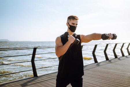 Handsome muscular man in protective mask, sports outfit doing functional exercises on seaside promenade.の写真素材