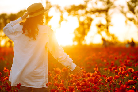 Young girl in hat posing in the poppy field. Portrait of curly girl walking in poppy field at sunset.の写真素材