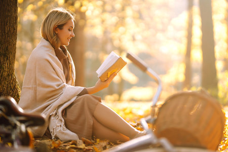 Beautiful young woman sitting on a fallen autumn leaves in a park, reading a book. relaxation.の写真素材