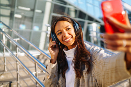 Optimistic woman wearing headphones and holding phone outdoors. Communication in social networks.の写真素材